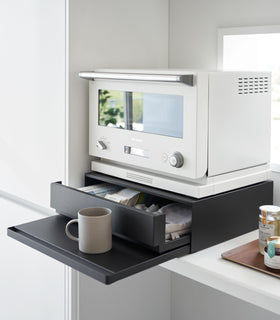 Diagonal view of the Countertop Drawer with Pull-Out Shelf by Yamazaki Home in black with the drawer and shelf pulled out. A mug sits on the shelf and a small microwave sits atop the unit. view 10