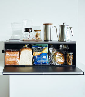 Frontal view of black Yamazaki Home Bread Box - Wide open with bread and snacks inside and coffee equipment on top view 12