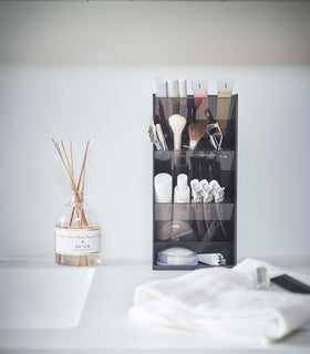 A front view of a black rectangular resin cosmetics organizer on a white bathroom counter. It has three deep adjustable black transparent trays that sit diagonally with matching dividers placed in the middle of each tray. view 12