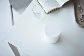 A bird’s-eye view of the corner of a white table. On the table is a cylinder-shaped white four-tier swivel accessory holder, a clear drinking glass, and an opened book. The accessory holder’s tiers and lid are swiveled closed so the inside contents cannot be seen, resembling a white cylinder object. view 16