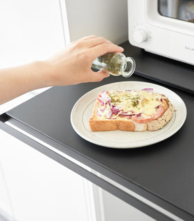 Close-up image of a hand seasoning food on a plate placed on the pulled out shelf of the Countertop Drawer with Pull-Out Shelf by Yamazaki Home in black. view 15