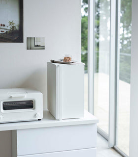 A vertical white metal breadbox is seen on a white kitchen counter next to a white microwave oven. The breadbox’s door is closed and has a long vertical handle. On top of the box is a folded towel and plastic container of cookies. Behind the counter is a white wall with two different sized postcards positioned diagonally from each other. view 15