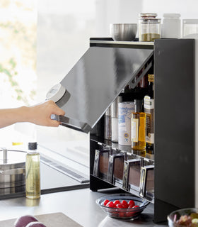 Image showing a hand holding the front panel of the Concealable Spice Rack by Yamazaki Home by the handle which is half open, revewling various condiments inside. view 12