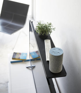 Aerial side view of black Compliant TV Shelf holding plant, smart home device, and remote behind TV by Yamazaki Home. view 9