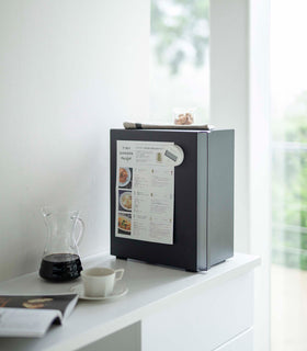 A profile view of a vertical black metal bread box sat on a white kitchen counter.  A long vertical handle is positioned on the left at the front of the box. A magnetic timer is holding up a recipe card on the side of the breadbox. A folded towel and plastic container of treats is laid on top. On the counter in front of the breadbox is a half-filled glass coffee pot and white coffee cup on a matching saucer. An out-of-focus opened magazine is seen next to the cup. view 25