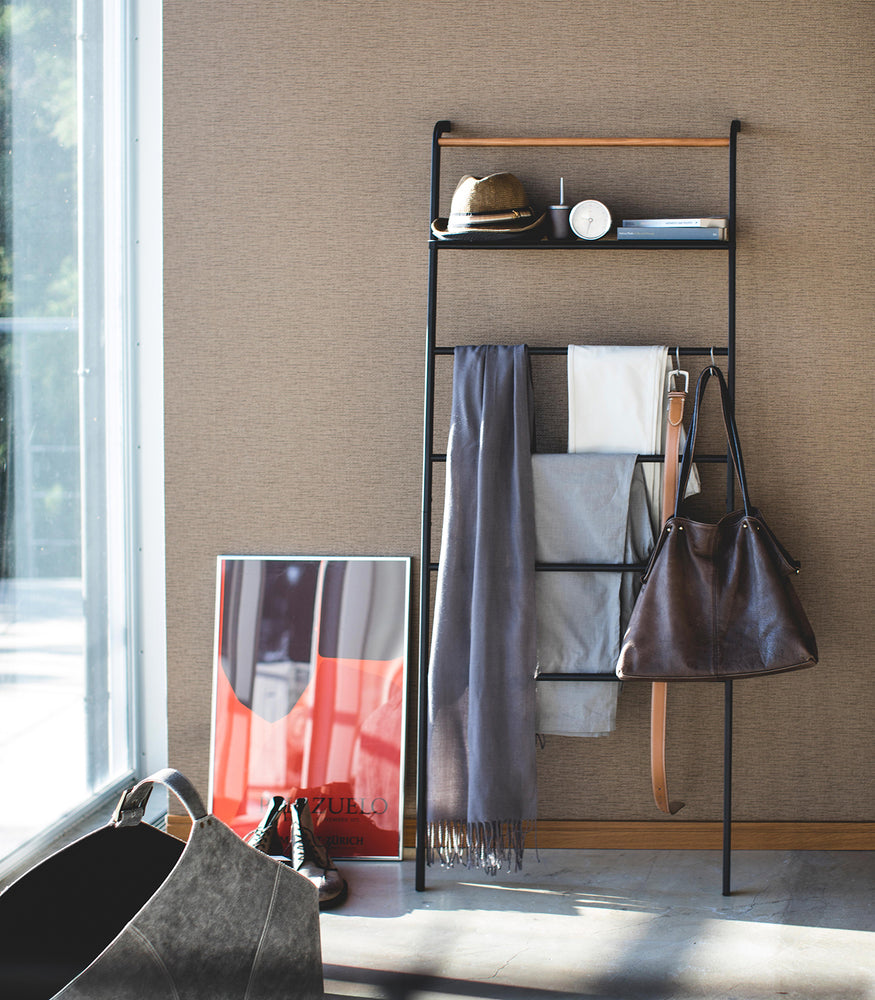 View 19 - Front view of black Leaning Ladder Rack with Shelf holding clothing items and purse in bedroom by Yamazaki Home.