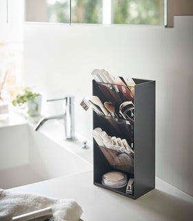 An angled view of a black rectangular resin cosmetics organizer on a white bathroom counter. It has an open face and top and three deep black adjustable transparent trays that sit diagonally with matching dividers placed in the middle of each tray. The bottom of the organizer has a raised lip and acts as additional storage. view 11