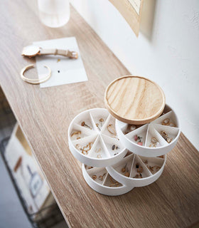 A bird’s eye view of the corner of a wooden console table. On the table is a cylinder-shaped white four-tier swivel accessory holder with a light-colored wooden lid. The accessory holder’s tiers and lid are swiveled opened so the inside contents can be seen. Each tier of the accessory holder is divided into seven separate compartments in a pie-chart fashion. view 9