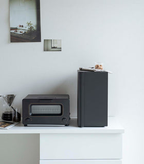 A vertical black metal breadbox is seen on a white kitchen counter next to a black microwave oven and a glass drip coffee pot with a dark liquid inside. The breadbox is closed and has a long vertical handle on the left of the box. On top of the box is a folded towel and plastic container of cookies. Behind the counter is a white wall with two different sized postcards positioned diagonally from each other. view 23