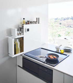 White Yamazaki Home Two-Tier Sliding Drawer Seasoning Rack with both tiers opened next to a stove top view 2