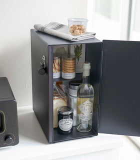 A vertical black metal breadbox is seen on a white kitchen counter next to a black microwave oven. The breadbox’s door is opened from the right and bread and other grains are seen inside the box. A magnetic stop is seen opposite the open door. A magnetic hook is attached to the side. On top of the box is a folded towel and plastic container of cookies. view 28