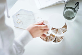 A bird’s-eye view of a hand reaching for something in a white four-tier swivel accessory holder on a white surface. view 20