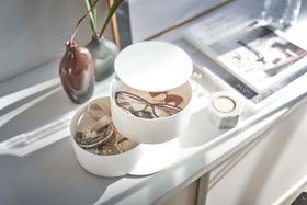 An angled bird’s-eye view of a two-tier white swivel accessory holder placed on a white dresser with two decorative vases and other décor. The accessory holder’s tiers and lid are swiveled opened so the inside contents can be seen. In the first tier are a pair of reading glasses, earrings, and other various jewelry. The second tier holds a watch and other jewelry. view 4