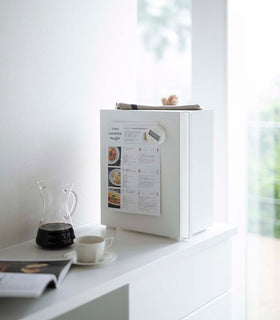 A profile view of a vertical white metal bread box sat on a white kitchen counter.  A long vertical handle is positioned on the left at the front of the box. A magnetic timer is holding up a recipe card on the side of the breadbox. A folded towel and plastic container of treats is laid on top. On the counter in front of the breadbox is a half-filled glass coffee pot and white coffee cup on a matching saucer. An out-of-focus opened magazine is seen next to the cup. view 18