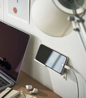 An iPhone with a charger cord plugged in is mounted on a wall above a desk setup. The screen is facing outward and the phone is held up on its side. view 2