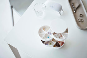 A bird’s-eye view of a white four-tier swivel accessory holder on a white surface. The accessory holder’s tiers and lid are swiveled opened so the inside contents can be seen, each tier is divided into seven individual compartments in a pie-shaped fashion. Each individual compartment of the tiers holds various beads and jewelry-making parts. In the background is an oval shaped catch-all plate with a pair of silver needle nose pliers and three rings. view 18