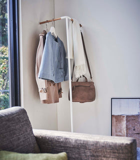 Side view of Yamazaki white Corner Leaning Coat Hanger in a the corner of a room with clothing hung on it view 4