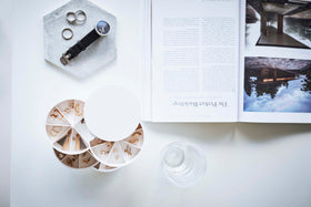 A birds-eye view of a white surface. On the surface is a cylinder-shaped white four-tier swivel accessory holder, a clear drinking glass, a geometric shaped catch-all plate and an upside-down opened book. A men’s watch and various rings are strewn on the catch-all dish. Each tier of the accessory holder is divided into seven separate compartments in a pie-chart fashion. Each compartment holds a set of earrings. view 19