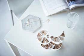 A bird’s-eye view of the corner of a white table. On the table are a white four-tier swivel accessory holder, a geometric shaped catch-all dish, a clear drinking glass, and an opened book. The accessory holder’s tiers and lid are swiveled opened so the inside contents can be seen, each tier has 7 individual compartments in a pie-shaped fashion. Each individual compartment of the tiers holds various earrings. view 15