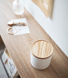 A bird’s eye view of the corner of a wooden console table. On the table is a cylinder-shaped white four-tier swivel accessory holder with a light-colored wooden lid. The accessory holder’s tiers and lid are swiveled closed so the inside contents are hidden away. To the left of the holder is a white paper with a gold watch and bracelet laid out on top. view 11