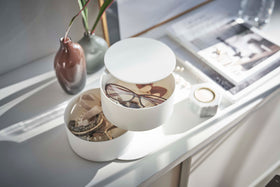 An angled bird’s-eye view of a two-tier white swivel accessory holder placed on a white dresser with two decorative vases and other décor in the background. The accessory holder’s tiers and lid are swiveled opened so the inside contents can be seen. In the first tier are a pair of reading glasses, earrings, and other various jewelry. The second tier holds a watch and an assortment of bracelets. view 8