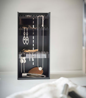 Sitting on top of a white bathroom counter is a black resin rectangle jewelry organizer. It has an open face and top with three transparent shelves with upward facing hooks along the edges, the last section of the rectangle is bare for added storage and has a small upward facing lip. view 12