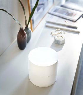 A profile view of a two-tier white swivel accessory holder on a white dresser with two decorative vases in the background along with other out-of-focus home décor. The accessory holder’s tiers and lid are closed so the inside contents can not be seen, resembling a white cylinder. view 3