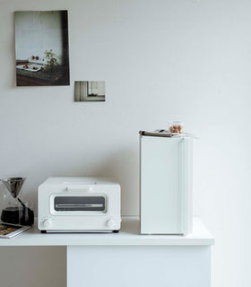 A vertical white metal breadbox with a long vertical handle on the left side is seen on a white kitchen counter next to a white microwave oven and a glass drip coffee pot with a dark liquid inside. On top of the box is a folded towel and plastic container of cookies. Behind the counter is a white wall with two different sized postcards positioned diagonally from each other. view 20