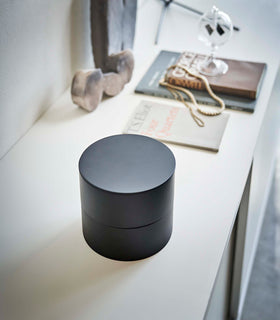 A profile view of a black two-tier swivel accessory holder on a white dresser. The accessory holder’s tiers and lid are closed so the inside contents cannot be seen, resembling a black cylinder. Out-of-focus in the background is a stone sculpture and a short stack of books with two gold chains strewn on top. Sunlight is coming from the right. view 11