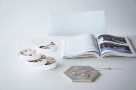 A front facing view of a white table with a white chair pushed in. On the surface of the table are a cylinder-shaped white four-tier swivel accessory holder, a clear drinking glass, a geometric shaped catch-all plate and an opened book facing the direction of the chair. view 17