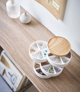 A bird’s-eye view of a wooden surface with a white four-tier swivel accessory holder with a light-colored wooden lid placed at the center. The accessory holder’s tiers and lid are swiveled opened so the inside contents can be seen, each tier is divided into seven individual compartments in a pie-shaped fashion. Each individual compartment of the tiers holds various beads and jewelry-making parts. view 12