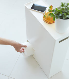 Aerial view of white Toilet Paper Stocker displaying phone, jar, and plant on top and dispensing a toilet paper roll by Yamazaki Home. view 4